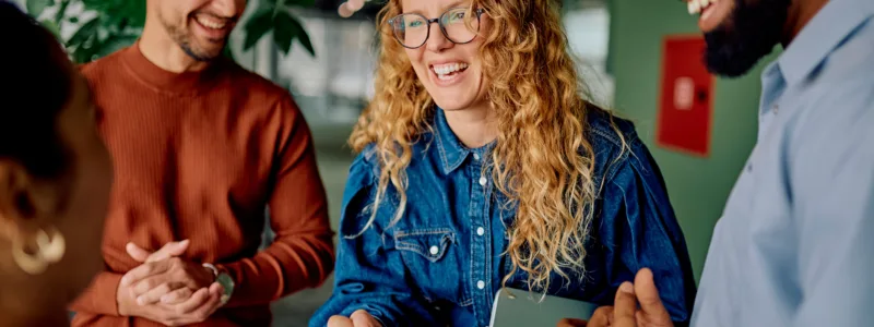 Four people stand together indoors, smiling and shaking hands, with one person holding a tablet. The group appears to be having a friendly conversation.