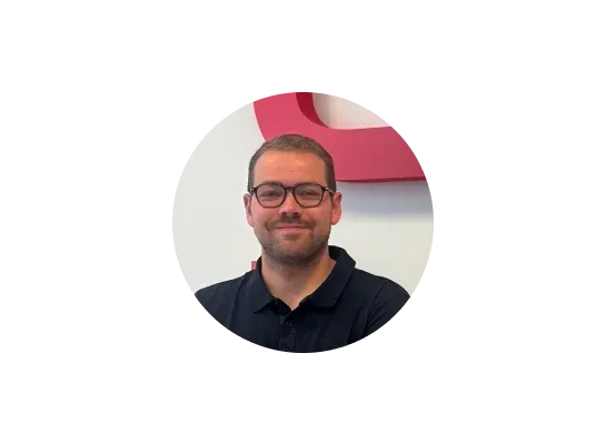 A man with short hair, glasses, and a beard, wearing a dark polo shirt, poses in front of a white wall with a red curved shape.