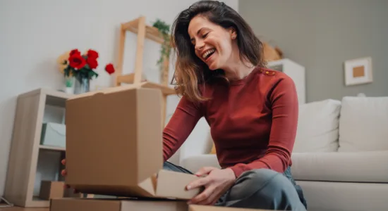 A woman sits on the floor in a living room, smiling as she opens a cardboard box, with other boxes stacked nearby.