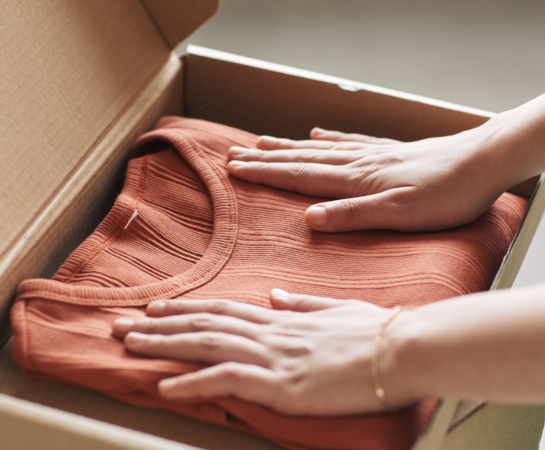 A folded orange shirt placed in an open cardboard box, with two hands gently packing it.