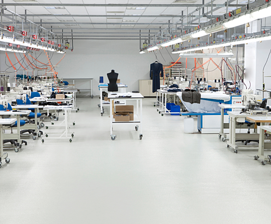 A spacious garment factory room with sewing machines and mannequins. Workstations are aligned in rows with overhead lighting and visible piping, creating an organized industrial look.
