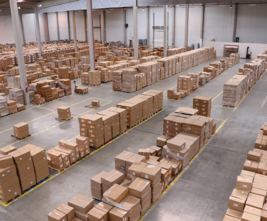 A large warehouse filled with organized stacks of cardboard boxes on pallets, lined up in rows. The warehouse has high ceilings and concrete floors.