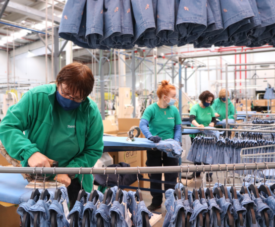 Workers in green uniforms and masks attend to jeans in a factory setting, with racks of denim garments hanging around them.
