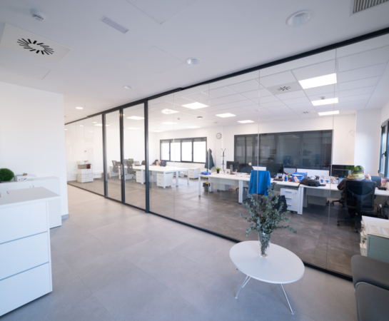 Modern office with glass partition, white desks, and chairs. A small table with a plant is in the foreground, and large windows let in natural light.