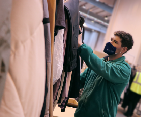 A person in a green jacket and face mask examines clothing on a rack in a large indoor space.