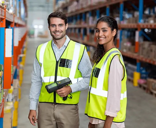 Two individuals wearing safety vests standing inside a warehouse.