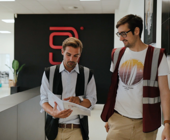 Two men wearing safety vests walk indoors, reviewing documents together near a black wall with a red logo.