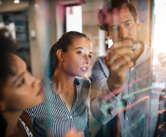 Three people stand behind a transparent board covered in colorful notes and diagrams, focusing intently as one person writes with a marker.