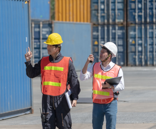 Two individuals wearing safety vests strolling near shipping containers in an industrial setting.