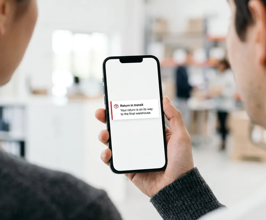 Two people look at a smartphone screen displaying a message that says, “Return in transit. Your return is on its way to the final warehouse.”.