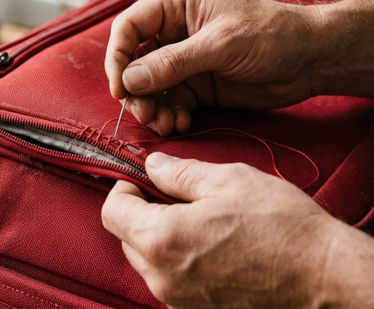 Close-up of hands using a needle and red thread to repair a tear near the zipper of a red fabric bag.