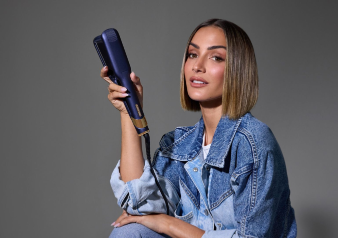 Person in denim jacket holding a hair straightener, sitting against a neutral background.