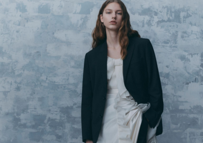 A person with long hair is wearing a dark blazer over a white dress, standing against a textured gray background.