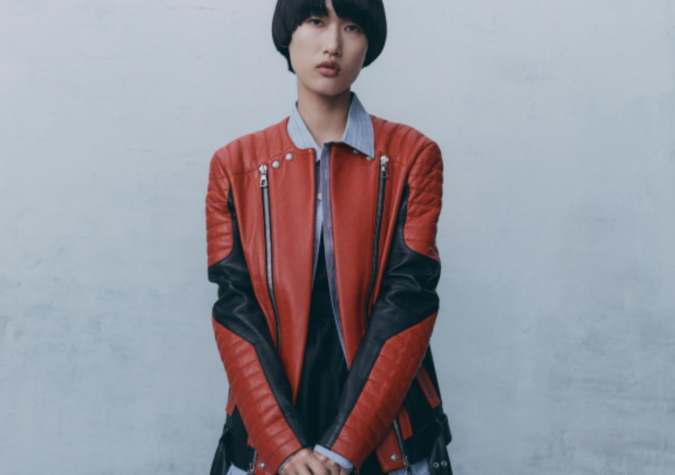 Person with short hair wearing a red and black leather jacket standing against a plain wall.