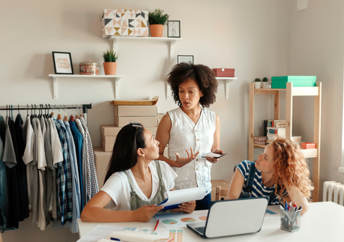 Three women are having a discussion in a room with clothing racks and shelves. One stands holding a phone, while the other two sit at a table with a laptop and papers.