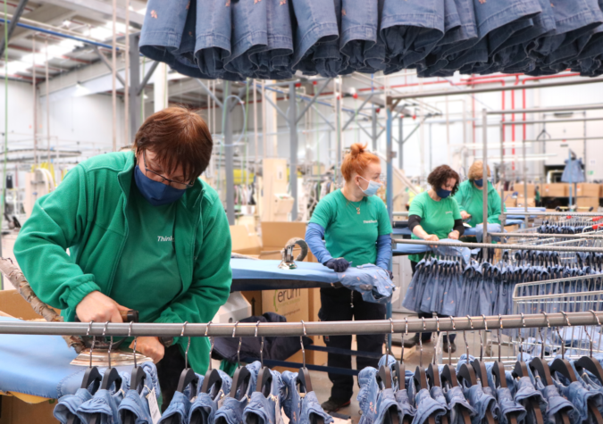 Workers in green shirts and masks operate in a factory, processing blue denim clothing items on racks. The setting is an industrial textile facility.