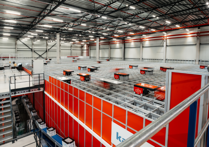Automated robots move along a grid system inside a large, modern warehouse with high ceilings and industrial lighting.