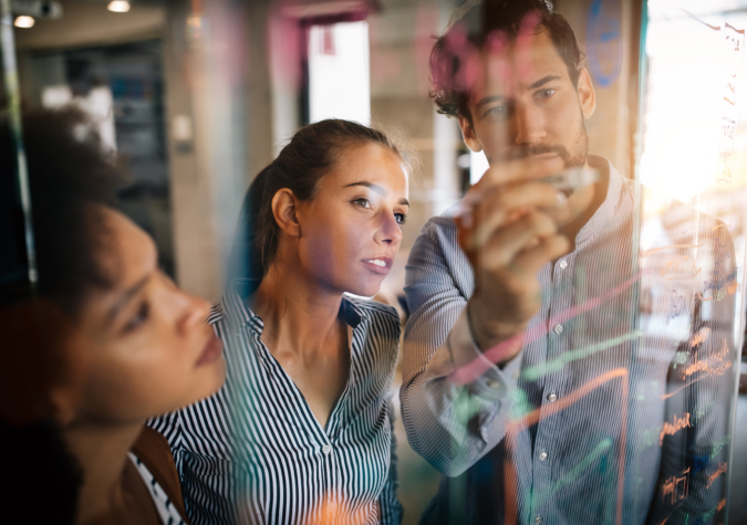 Three people stand behind a transparent board covered in colorful notes and diagrams, focusing intently as one person writes with a marker.