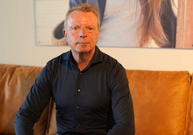 A middle-aged man in a dark shirt sits on a tan couch indoors, facing the camera with a neutral expression. A blurred photo is on the wall behind him.