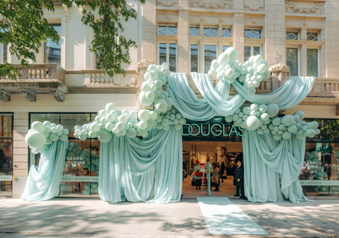 Storefront decorated with large clusters of light blue balloons and draped fabric, with “Douglas” sign above the entrance and people visible inside the store.