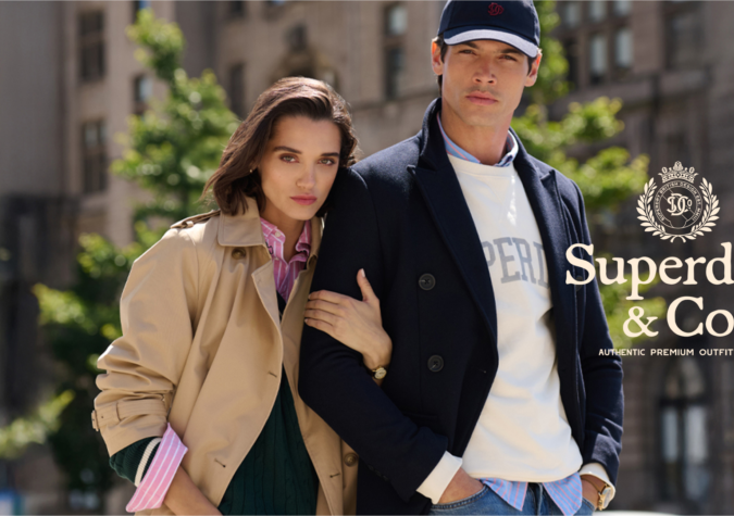 A woman and a man in stylish Superdry & Co outfits pose outdoors in an urban setting with blurred buildings and trees in the background.