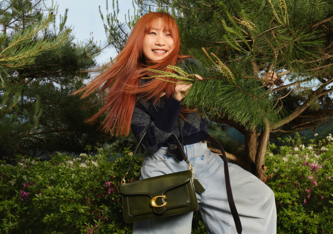 A woman with long red hair stands outdoors by a pine tree, smiling, wearing a navy sweater, light blue jeans, and carrying a green handbag.