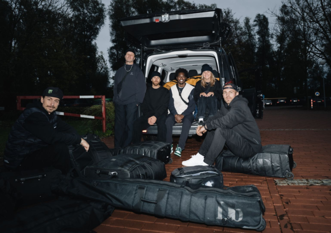 Six people sit and squat near an open van trunk, surrounded by multiple large black bags, in an outdoor setting with trees and red brick pavement.