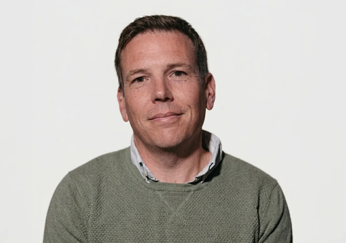 A man with short brown hair, wearing a green sweater over a collared shirt, sits in front of a plain white background, looking at the camera.