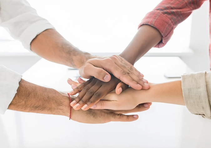 Four people stack their hands together in a gesture of teamwork against a bright background.