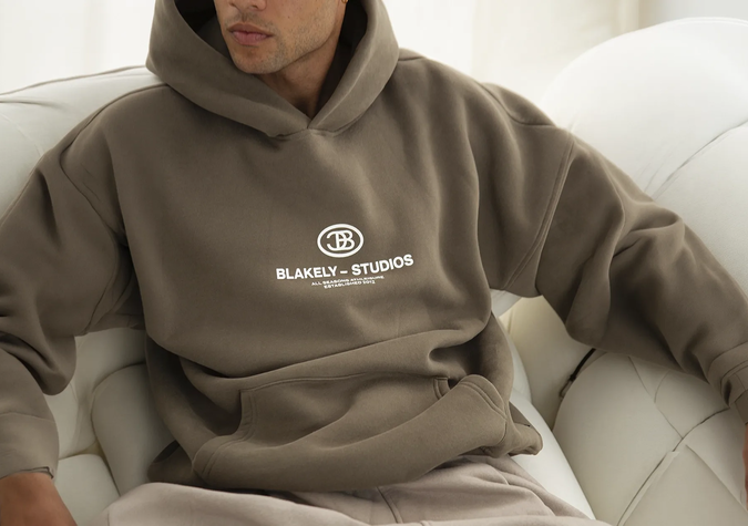 A person wearing a taupe Blakely Studios hoodie is seated on a white couch, with one arm resting on the back of the couch.