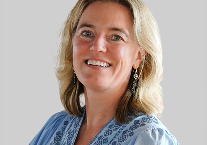 A woman with shoulder-length blonde hair, wearing a light blue patterned blouse and dangling earrings, smiles at the camera against a plain light background.