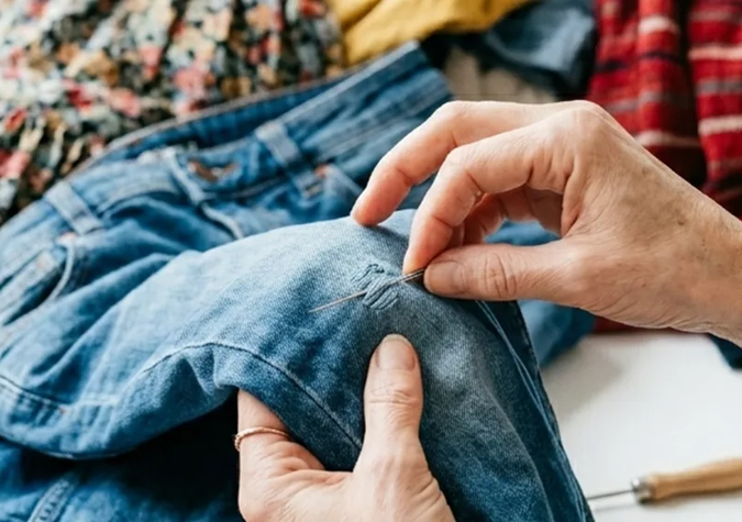 Hands repairing a tear in blue denim jeans with a needle and thread, with various clothing items in the background.