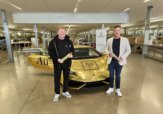 Two men stand in front of a gold car with "Au Vodka" branding inside a modern, well-lit building, each holding a rolled-up item while smiling at the camera.