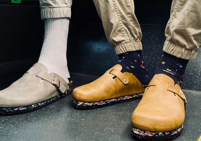Two people wearing slip-on clogs; one has beige shoes with white socks and tan pants, the other has tan shoes with dark patterned socks and khaki pants, standing on a speckled floor.