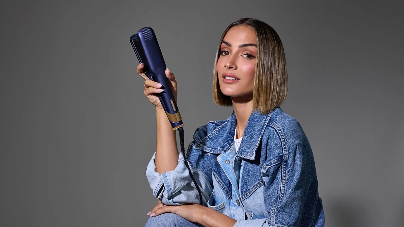 Person in denim jacket holding a hair straightener, sitting against a neutral background.