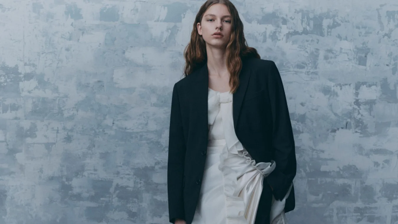 A person with long hair is wearing a dark blazer over a white dress, standing against a textured gray background.