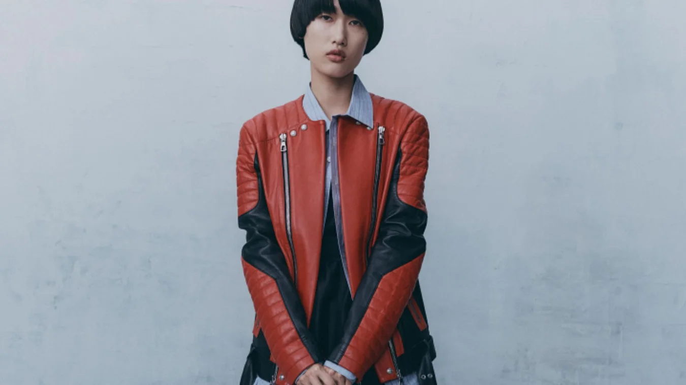 Person with short hair wearing a red and black leather jacket standing against a plain wall.