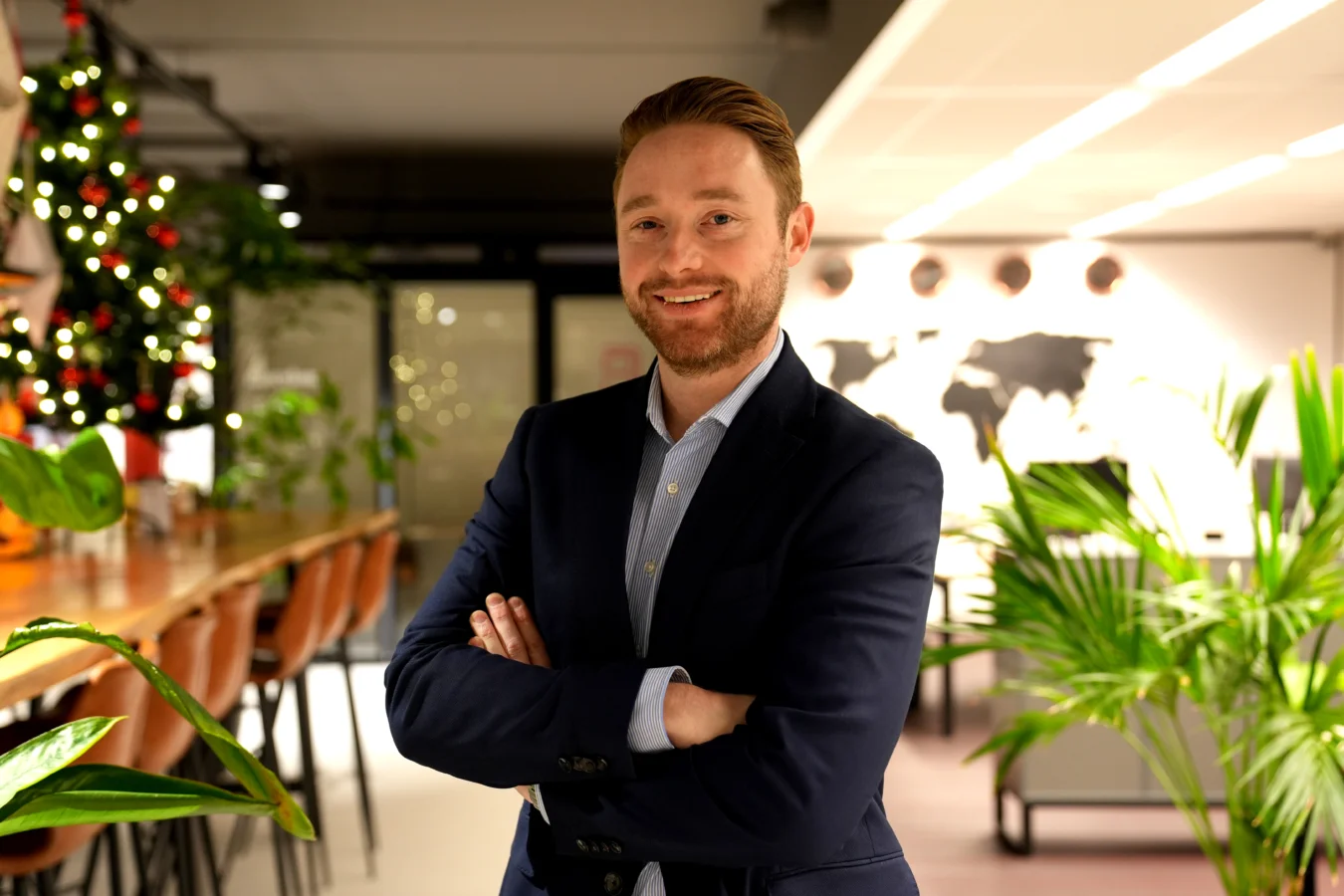A man in a blue blazer stands with arms crossed in a modern office. There is a world map on the wall and a decorated Christmas tree to the left.