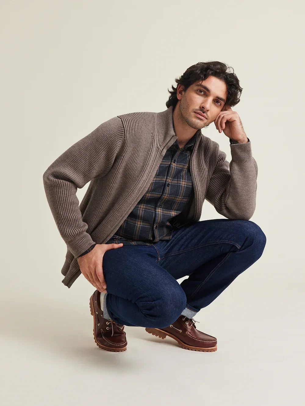 A person squatting, wearing a brown sweater, plaid shirt, blue jeans, and brown shoes, with a neutral expression, against a plain background.