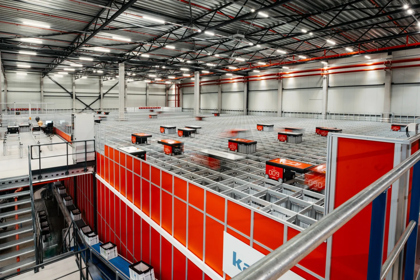 Automated robots move along a grid system inside a large, modern warehouse with high ceilings and industrial lighting.