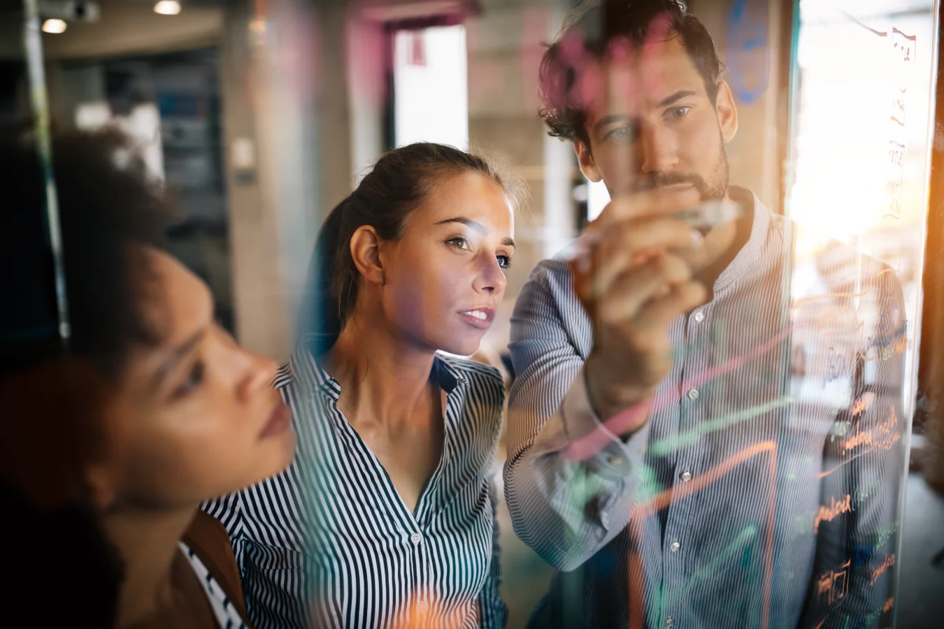 Three people stand behind a transparent board covered in colorful notes and diagrams, focusing intently as one person writes with a marker.