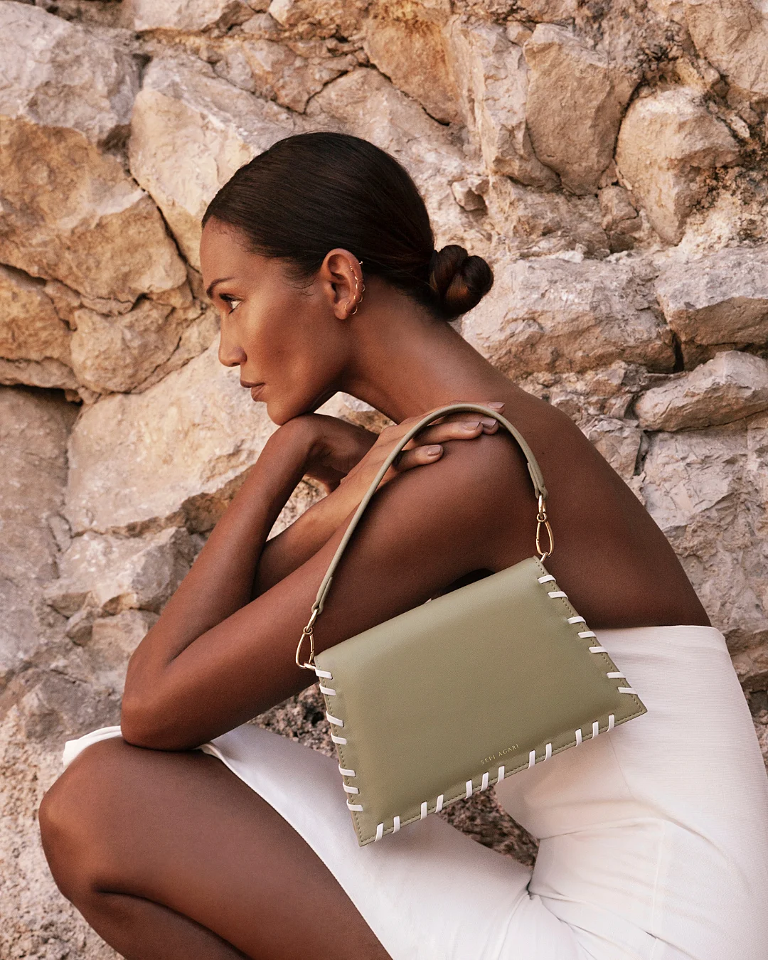 A woman with a sleek bun sits sideways against a stone wall, wearing a white dress and holding a sage green handbag with white stitching.