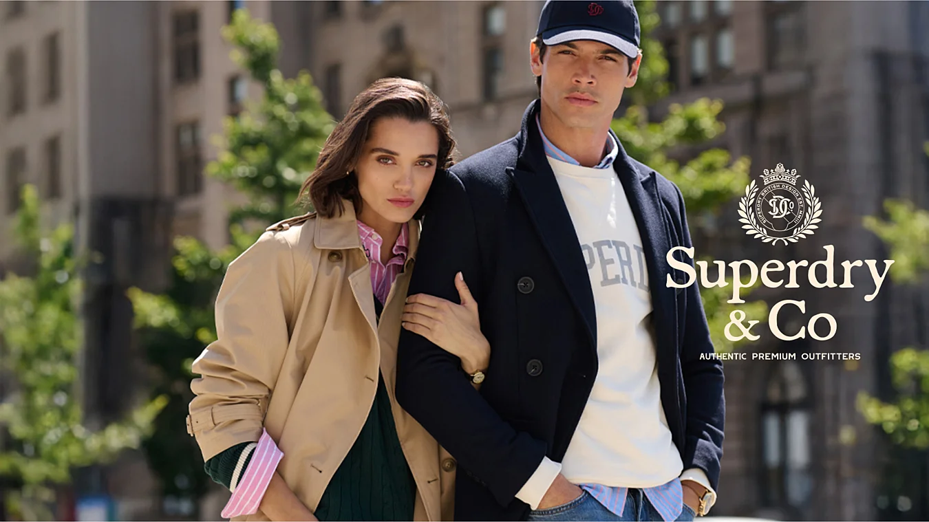A woman and a man in stylish Superdry & Co outfits pose outdoors in an urban setting with blurred buildings and trees in the background.