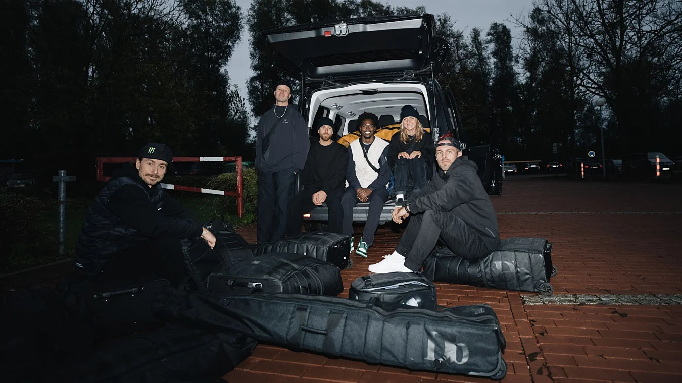 Six people sit and squat near an open van trunk, surrounded by multiple large black bags, in an outdoor setting with trees and red brick pavement.