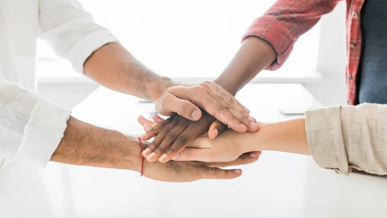Four people stack their hands together in a gesture of teamwork against a bright background.