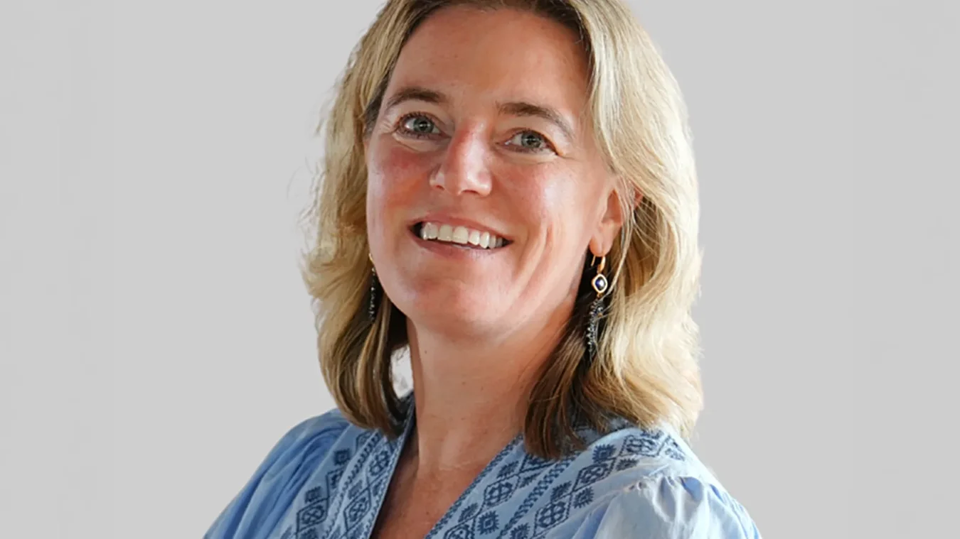 A woman with shoulder-length blonde hair, wearing a light blue patterned blouse and dangling earrings, smiles at the camera against a plain light background.