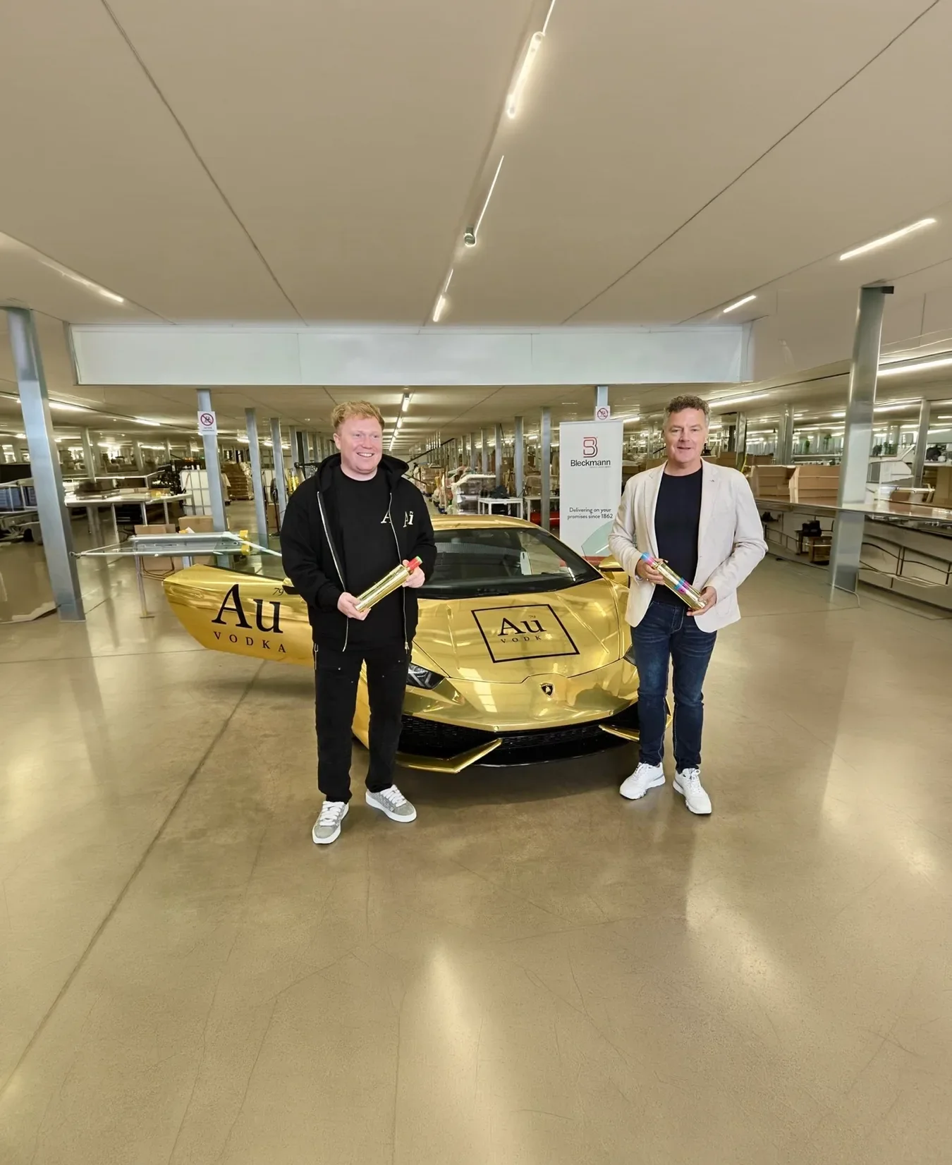 Two men stand in front of a gold car with "Au Vodka" branding inside a modern, well-lit building, each holding a rolled-up item while smiling at the camera.