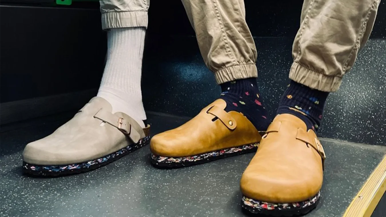 Two people wearing slip-on clogs; one has beige shoes with white socks and tan pants, the other has tan shoes with dark patterned socks and khaki pants, standing on a speckled floor.
