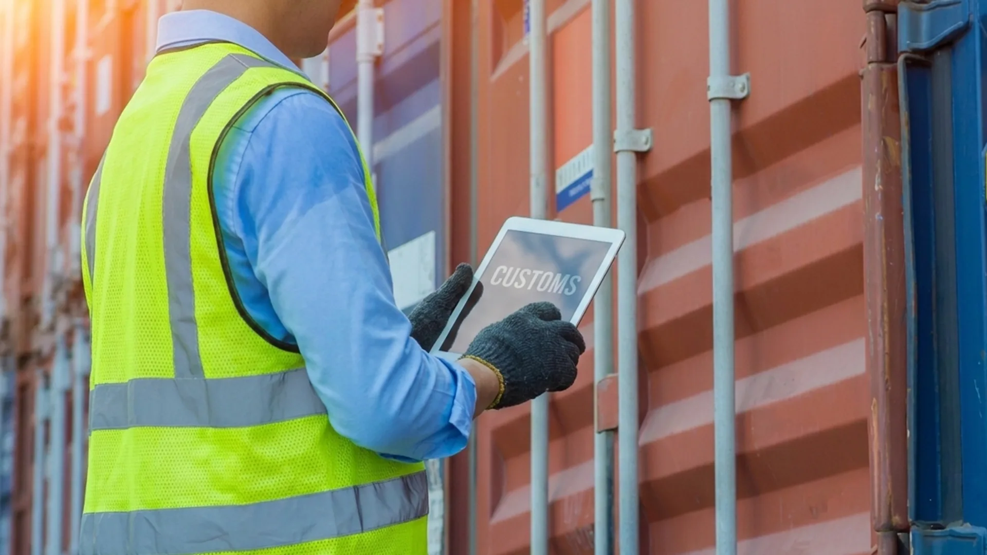 A person wearing a safety vest and gloves holds a tablet displaying "CUSTOMS" while inspecting a shipping container.
