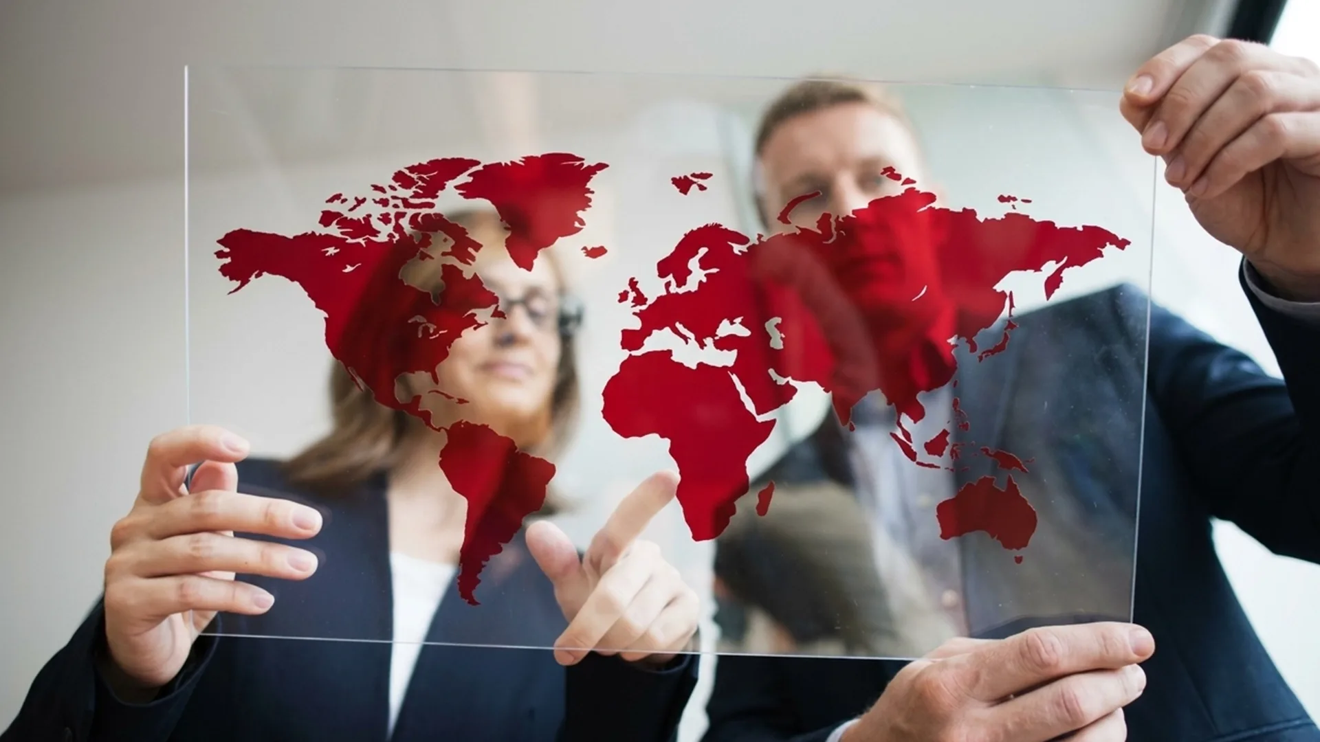 Two people in business attire hold a transparent panel with a red world map, pointing at different locations.
